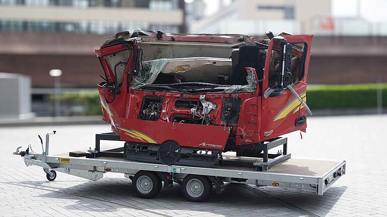 Stark deformiertes, rotes Fahrerhaus eines Lkw, das nach einem schweren Unfall auf einem Anhänger zu Ausstellungszwecken transportiert wird. Die Kabine ist stark eingedrückt, Scheiben sind zerbrochen und Verformungen deutlich sichtbar.