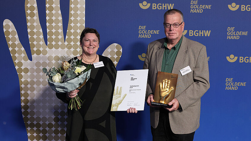 Eine Frau mit Blumenstrauß und ein Mann mit Urkunde und Goldene-Hand-Trophäe stehen vor der BGHW-Fotowand und lächeln in die Kamera.