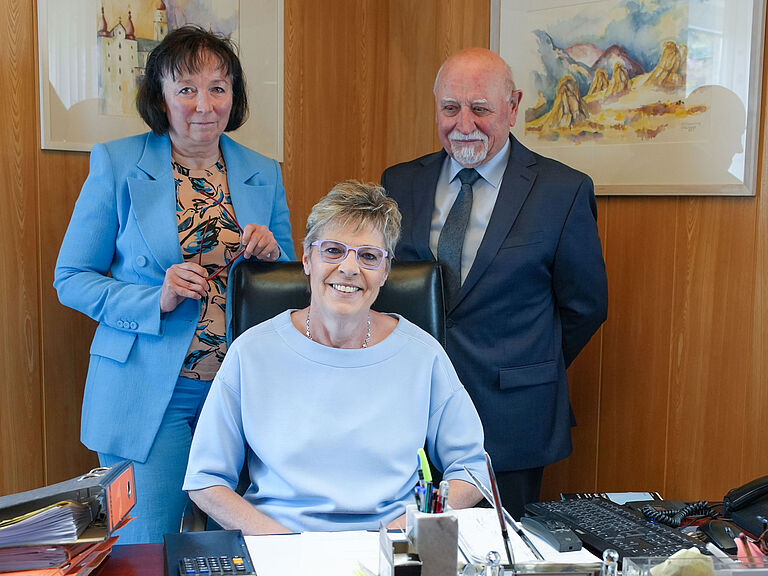 Die Rechtsanwälte Sylvia und Franz Grasser - Tochter und Vater - stehen hinter ihrer Mitarbeiterin Ute Grötznger, die am Schreitisch ihres Chefs sitzt. Alle schauen freundlich in die Kamera.