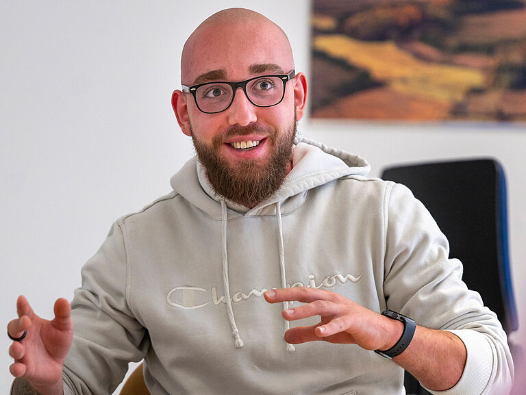 Moritz Sontheimer sitzt am Tisch und spricht gestikulierend. Er schaut dabei freundlich sein Gegenüber an. 