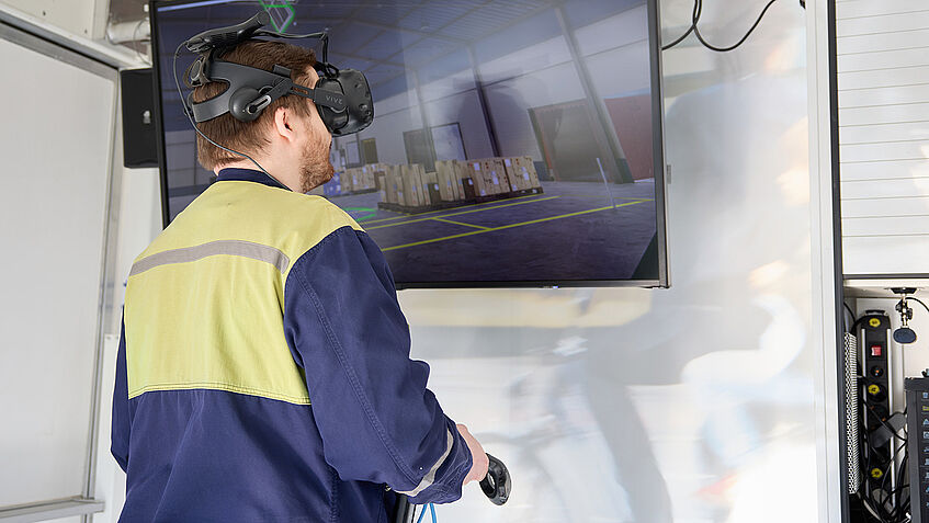 Ein Mann mit Sicherheitsjacke trägt eine VR-Brille, hält einen Joystick in der Hand und über ihm hängt ein Monitor.