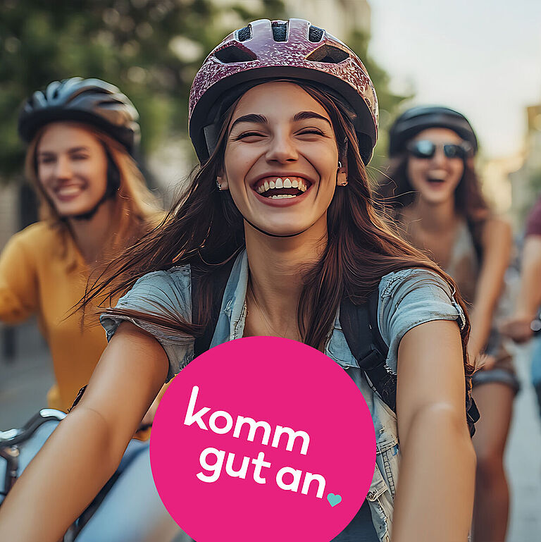Gruppe junger Menschen fährt lachend mit Fahrrad durch eine sonnige Innenstadt. Alle tragen Fahrradhelme. Im Vordergrund befindet sich ein pinkfarbener Kreis mit der Aufschrift „komm gut an“.