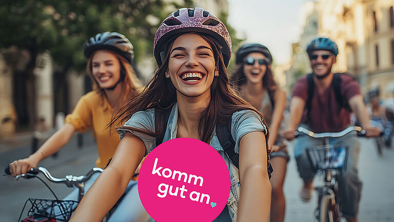Gruppe junger Menschen fährt lachend mit Fahrrad durch eine sonnige Innenstadt. Alle tragen Fahrradhelme. Im Vordergrund befindet sich ein pinkfarbener Kreis mit der Aufschrift „komm gut an“.