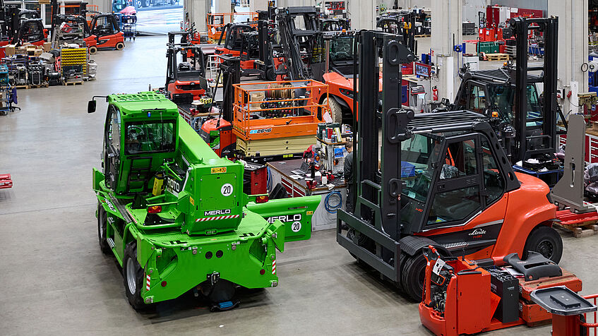 Blick in eine große Werkhalle mit mehreren Gabelstaplern und Arbeitsbereichen, in denen Personen an Fahrzeugen arbeiten.