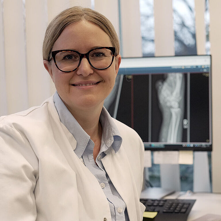Dr. Wenger schaut freundlich in die Kamera. Sie trägt eine Brille, einen weißen Kittel. Auf dem PC-Bildschirm im Hintergrund ist ein Röntgenbild zu sehen.