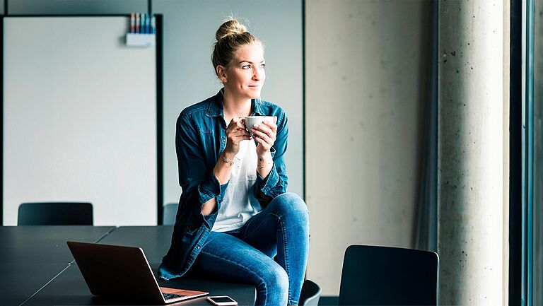 Eine Frau sitzt in einem Büro am Fenster, schaut hinaus und trinkt einen Kaffee