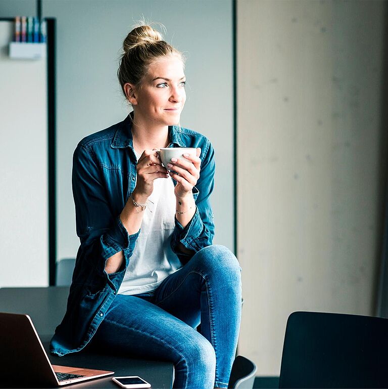 Eine Frau sitzt in einem Büro am Fenster, schaut hinaus und trinkt einen Kaffee