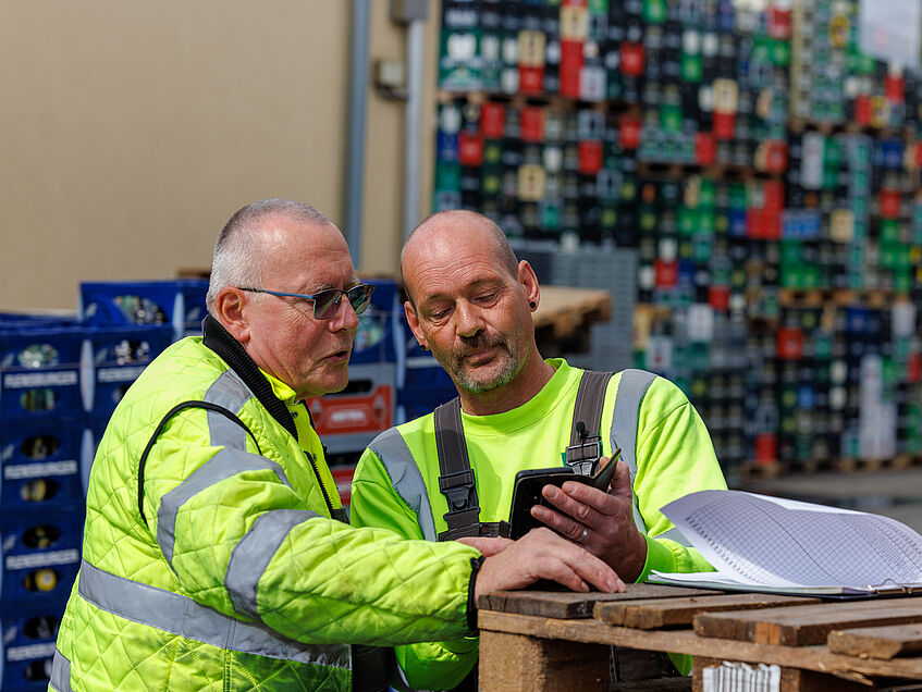 Dirk Boßmeyer und ein Kollege bei Getränke Essmann schauen auf sein Handy. Beide tragen gelbe Warnjacken. Im Hintergrund ist das Getränkelager.