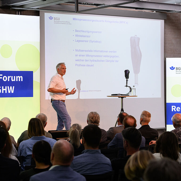 Dieses Bild zeigt Dr. Sebastian Benner, Sektionsleiter Technische Orthopädie, der BG Unfallklinik Frankfurt. Er spricht gestikulierend auf der Bühne vor Teilnehmenden des BGHW-Reha-Forums . Eine Beinprothese steht vor ihm auf der Bühne. Er spricht über Prothesentechnik und ihre Auswirkungen für Spitzensportler im Amputiertensport. 