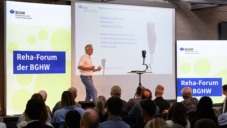 Dieses Bild zeigt Dr. Sebastian Benner, Sektionsleiter Technische Orthopädie, der BG Unfallklinik Frankfurt. Er spricht gestikulierend auf der Bühne vor Teilnehmenden des BGHW-Reha-Forums . Eine Beinprothese steht vor ihm auf der Bühne. Er spricht über Prothesentechnik und ihre Auswirkungen für Spitzensportler im Amputiertensport.
