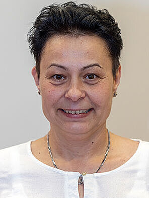 Porträt einer Frau mit kurzem dunklem Haar in weißer Bluse, frontal blickend, mit Namensschild. Es handelt sich um Jaqueline Riedel, ehrenamtliches Mitglied der BGHW-Selbstverwaltung für die Versichertenseite.