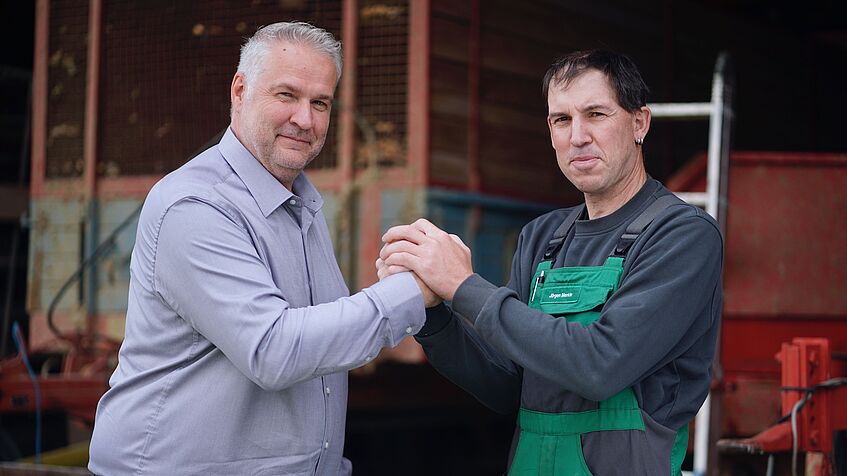 Zwei Männer schütteln sich die Hand, im Hintergrund Bauernhof-Ambiente.