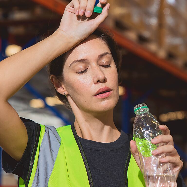 Eine Frau mit Warnweste und Wasserflasche in der Hand steht in einer Lagerhalle und wendet das Gesicht mit geschlossenen Augen in die Sonne, wobei sie sich mit der anderen Hand den Schweiß von der Stirn wischt.