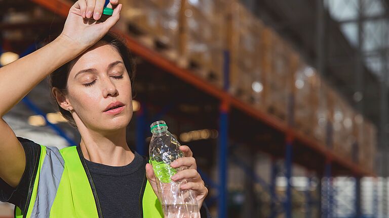 Eine Frau mit Warnweste und Wasserflasche in der Hand steht in einer Lagerhalle und wendet das Gesicht mit geschlossenen Augen in die Sonne, wobei sie sich mit der anderen Hand den Schweiß von der Stirn wischt.
