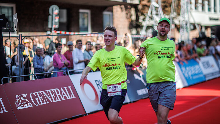 Zwei Menschen laufen in einem Staffellauf Hand in Hand. Einer von ihnen ist beeinträchtigt. Der Läufer rechts i Bild hat einen fehlgebildeten, kurzen Arm. Die Hand hält sein Laufpartner. Ober der andere Arm fehlt oder hinter dem Rücken ist, sieht man auf dem Bild nicht. Am Rand der abgesperrten Straße stehen viele Zuschauer. 