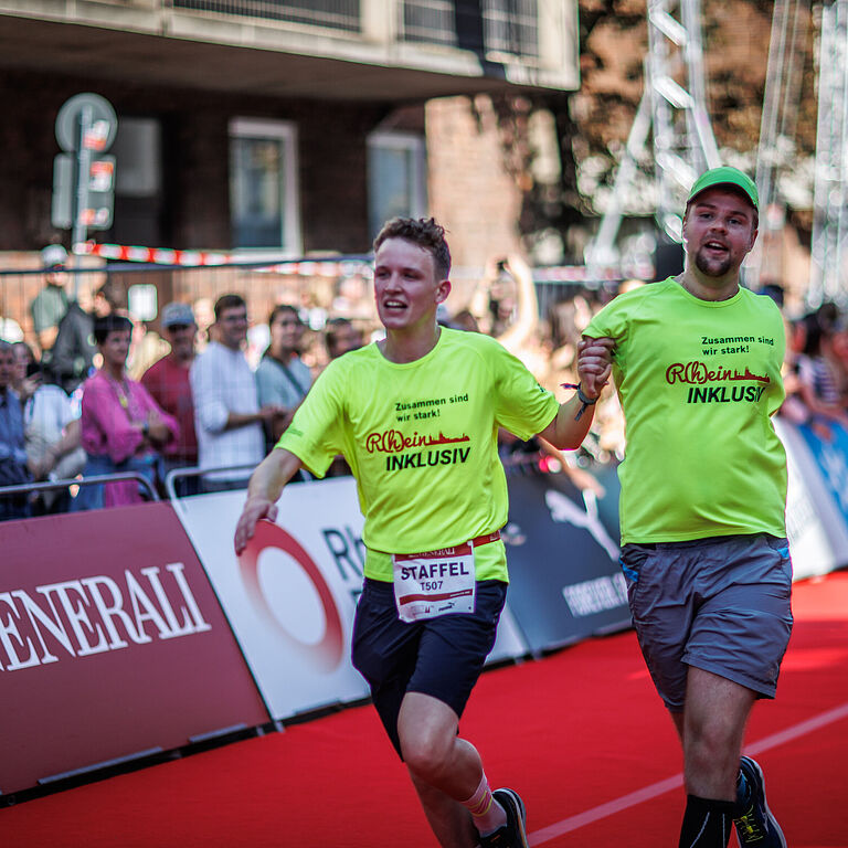 Zwei Menschen laufen in einem Staffellauf Hand in Hand. Einer von ihnen ist beeinträchtigt. Der Läufer rechts i Bild hat einen fehlgebildeten, kurzen Arm. Die Hand hält sein Laufpartner. Ober der andere Arm fehlt oder hinter dem Rücken ist, sieht man auf dem Bild nicht. Am Rand der abgesperrten Straße stehen viele Zuschauer. 