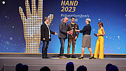 Blick auf die Bühne zur Präventionspreisverleihung: Links stehen drei Männer der Witt-Gruppe, die stolz die GOLDENE HAND von Silke Mayer-Seidler, Vorsitzende der Vertreterversammlung, entgegennehmen. Rechts im Bild steht Tina Babbel.