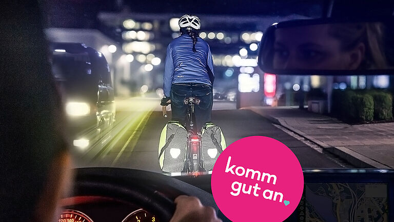 Eine Autofahrerin fährt nachts durch eine Stadtstraße und folgt einem Radfahrer mit Helm, reflektierender Jacke und leuchtenden Fahrradtaschen. Die Szene wird aus der Perspektive des Autos gezeigt. Im Rückspiegel ist das Gesicht der Fahrerin zu sehen. Im Vordergrund befinden sich das Lenkrad und ein Navigationsgerät. Auf dem Bild steht der Schriftzug „komm gut an“ in einem pinkfarbenen Kreis.