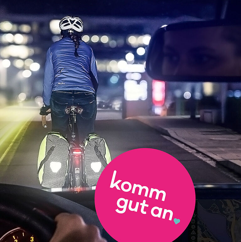 Eine Autofahrerin fährt nachts durch eine Stadtstraße und folgt einem Radfahrer mit Helm, reflektierender Jacke und leuchtenden Fahrradtaschen. Die Szene wird aus der Perspektive des Autos gezeigt. Im Rückspiegel ist das Gesicht der Fahrerin zu sehen. Im Vordergrund befinden sich das Lenkrad und ein Navigationsgerät. Auf dem Bild steht der Schriftzug „komm gut an“ in einem pinkfarbenen Kreis.
