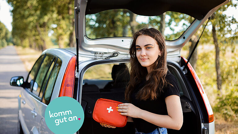 Das Bild zeigt eine Frau mit Kfz-Verbandkasten vor einem Kofferraum.
