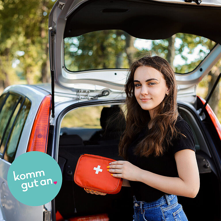 Das Bild zeigt eine Frau mit Kfz-Verbandkasten vor einem Kofferraum.