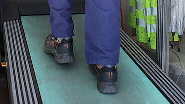 Man sieht von hinten die Beine einer Person in einer blauen Arbeitshose und schwarzen Sicherheitsschuhen, die auf einem Laufband geht.