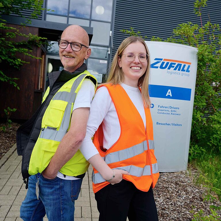 Zwei Personen in Warnwesten stehen Rücken an Rücken vor einem Firmengebäude. Im Hintergrund ist ein Schild der Zufall Logistics Group zu sehen.