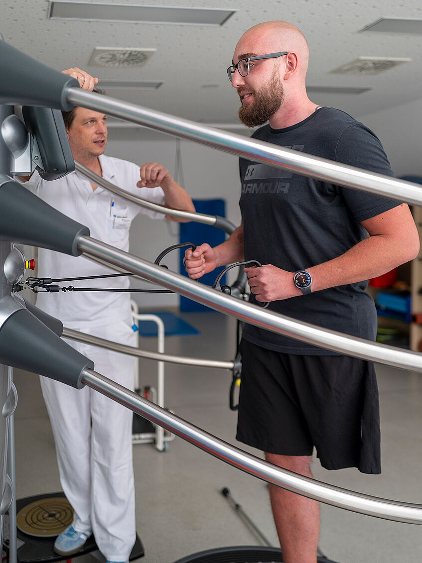 Moritz Sontheimer beim Physiotraining auf einer runden Platte. Links neben ihm: Haltestangen aus Metall, rechts der Physiotherapeut.