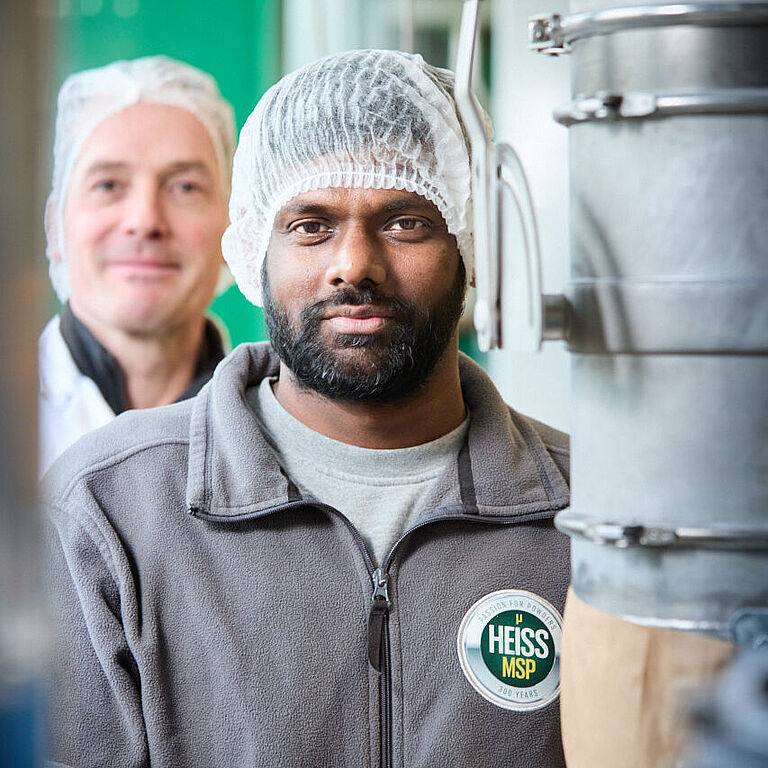 Zwei Männer stehen hintereinander und schauen zwischen zwei Metallrohren durch in die Kamera. Beide tragen weiße Hauben. Der Mann vorne hat schwarze Haare und einen Vollbart. 