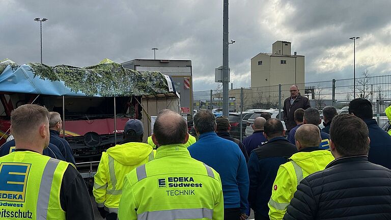 Edeka-Mitarbeiter stehen vor dem LKW-Exponat mit der zerquetschten Fahrerkabine und hören Dieter Schäfer zu