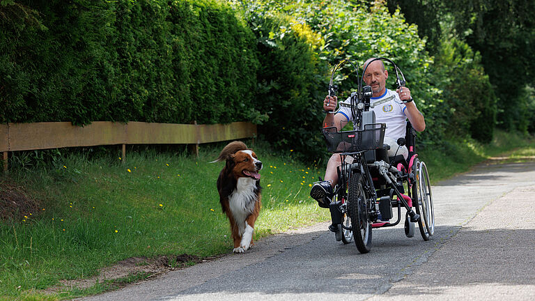 Dirk Boßmeyer trainiert mit seinem Hand-Bike, ein Rollstuhl-Zuggerät, das durch die Muskelkraft der Arme angetrieben wird. Der Schotterweg führt an einer grünen Hecke vorbei. Sein Hund begleitet ihn.