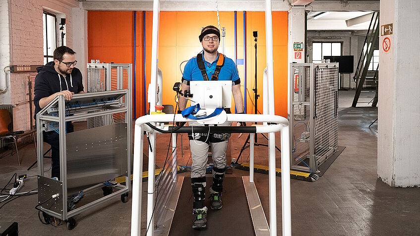 Blick in einen Trainingsraum, wo ein Mann, der verkabelt ist, auf einem Laufband geht. Ein Person an der Seite schaut zu.