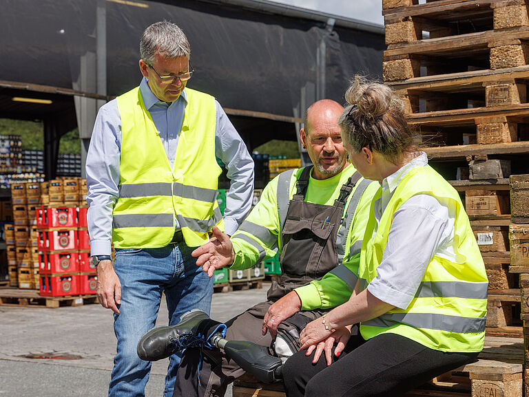 Dirk Boßmeyer sitzt im Getränkelager auf einem Stapel Paletten. Links und rechts von ihm die BGHW-Hilfsmittelbeauftragte Stephanie Wiebensohn und Reha-Berater Frank Heidemann. Beide tragen gelbe Warnwesten.