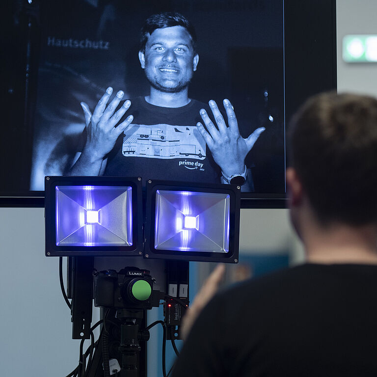 Von hinten ist Niklas Schrage zu sehen, der vor der UV-Kamera und einem Bildschirm sitzt. Er hält seine Hände in die Luft.