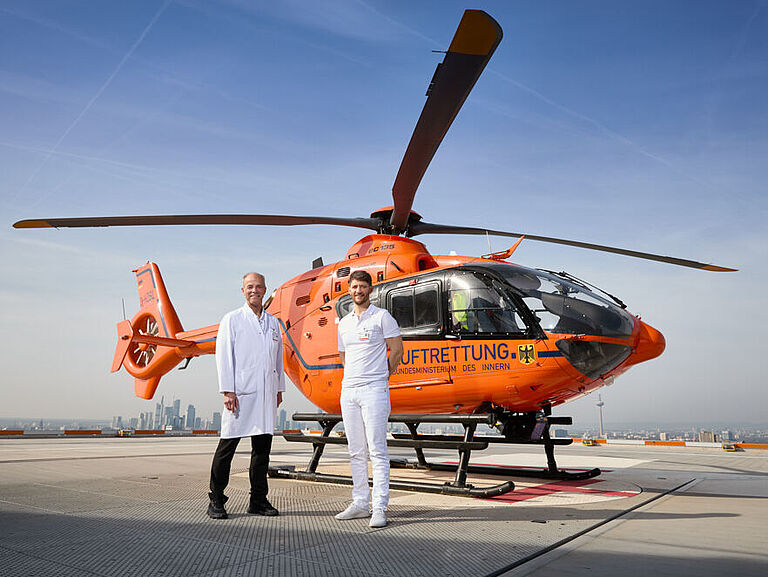 Der leuchtend rote Rettungshubschrauber der BG Klinik steht auf dem obersten Steockwerk. Davor stehen zwei Ärzte in weißen Kitteln. Im Hintergrund: die Frankfurter Skyline. 