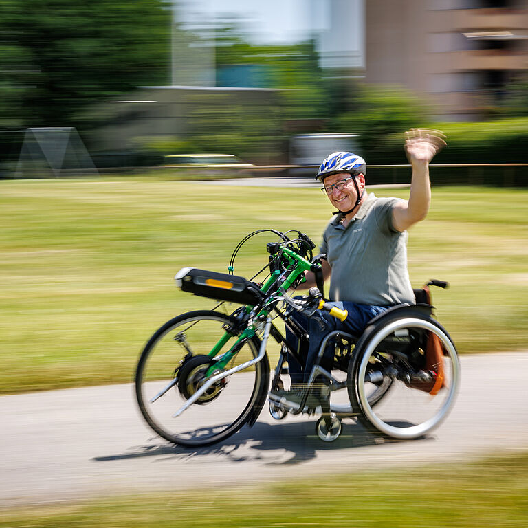 Axel Wisgalla fährt mit seinem Rollstuhl über einen asphaltierten Wirtschaftsweg. Er trägt einen Fahrradhelm und winkt lachend mit der linken Hand in die Kamera. Die Sonne lacht auch. Er fährt schnell.