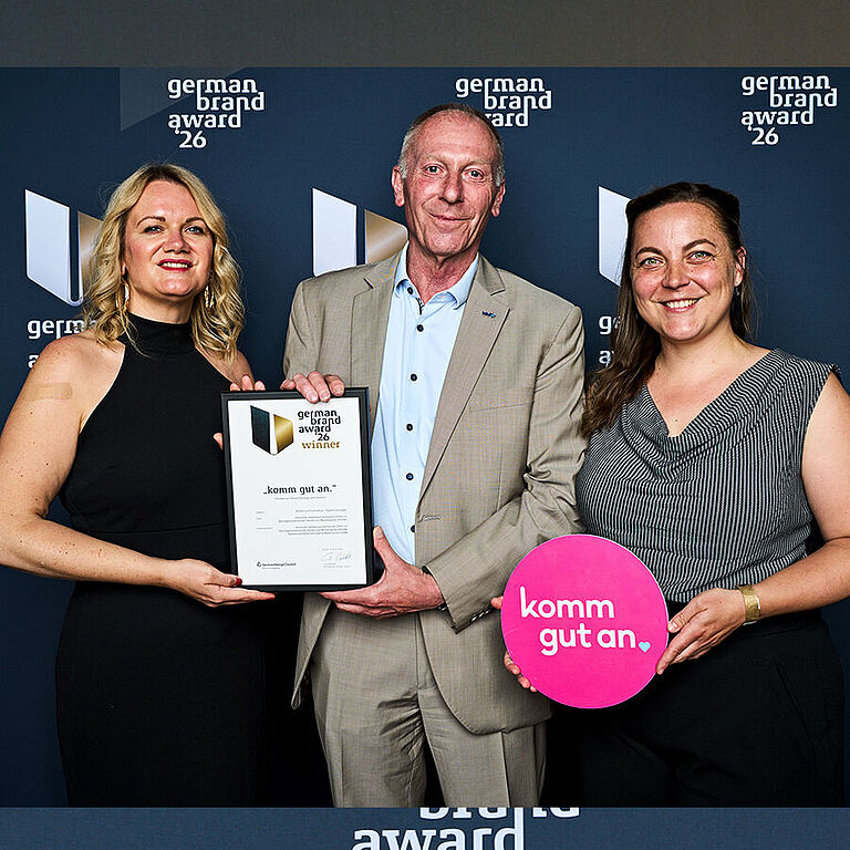 zwei Frauen und ein Mann präsentieren eine gerahmte Urkunde und ein Schild mit dem Titel "komm gut an"