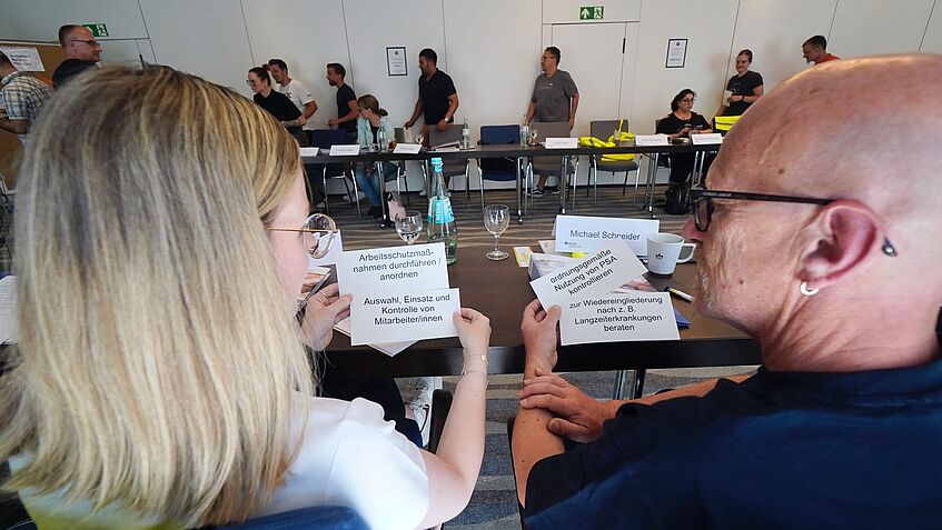 Zwei Personen sitzen nebeneinander und halten jeweils Kärtchen mit Text in den Händen. Auf den Karten stehen Aufgaben wie „Arbeitsschutzmaßnahmen durchführen/anordnen“, „Auswahl, Einsatz und Kontrolle von Mitarbeiter/innen“ und „ordnungsgemäße Nutzung von PSA kontrollieren“. Im Hintergrund sind weitere Seminarteilnehmende im Raum zu sehen.