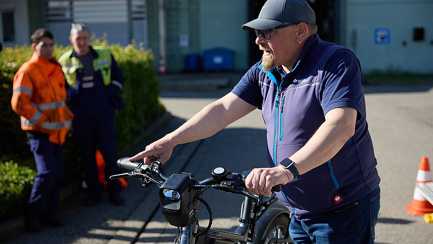 Ein Mann mit Kappi schiebt ein E-Bike, zwei Personen in Sicherheitskleidung schauen zu.