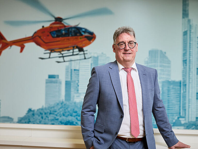 Martin Kögler, BGHW-Referent Teilhabe und stellvertretender Dezernatsleiter Rehabilitation und Leistungen, steht an ein Geländer angelehnt. Er trägt ein blaues Jackett und eine rot-weiße Krawatte und schaut in die Kamera. Im Hintergrund sind die Skyline von Frankfurt und ein Rettungshubschrauber auf die Wand gemalt. 