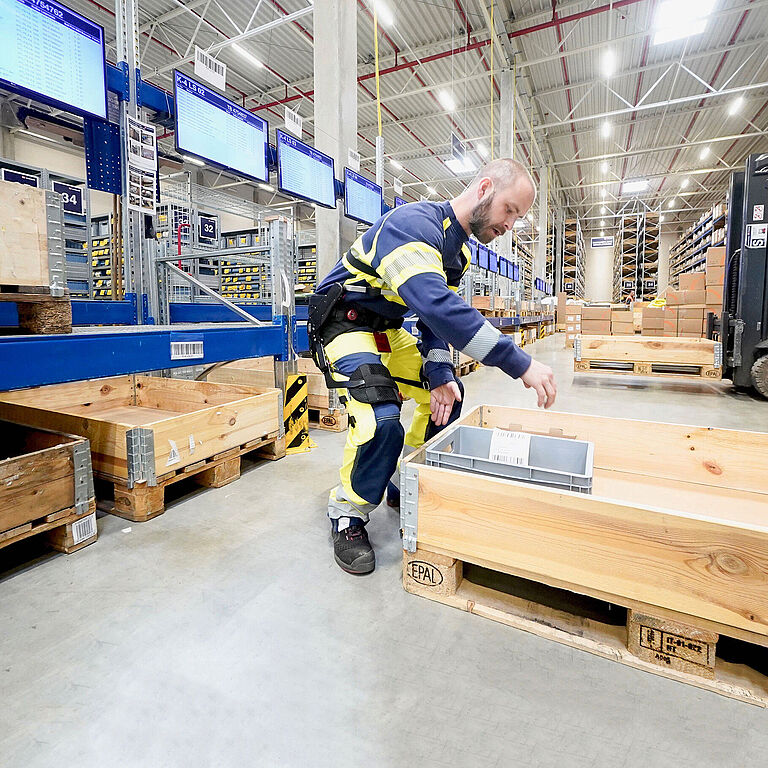 Ein Beschäftigter trägt ein Exoskelett und hebt eine Last in einem Lager an