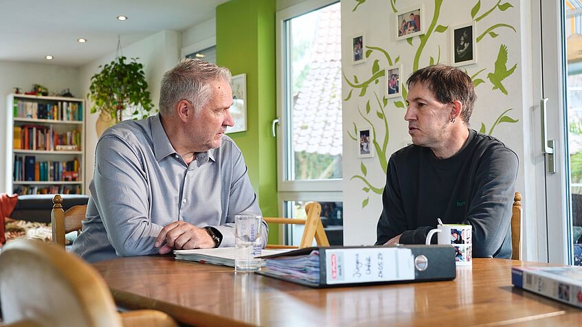 Zwei Männer im Gespräch an einem Tisch, auf dem Tisch liegt ein Aktenordner