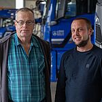 Zwei Männer stehen nebeneinander in einer Fahrzeughalle und schauen in die Kamera. Im Hintergrund stehen parkende Lkw. 