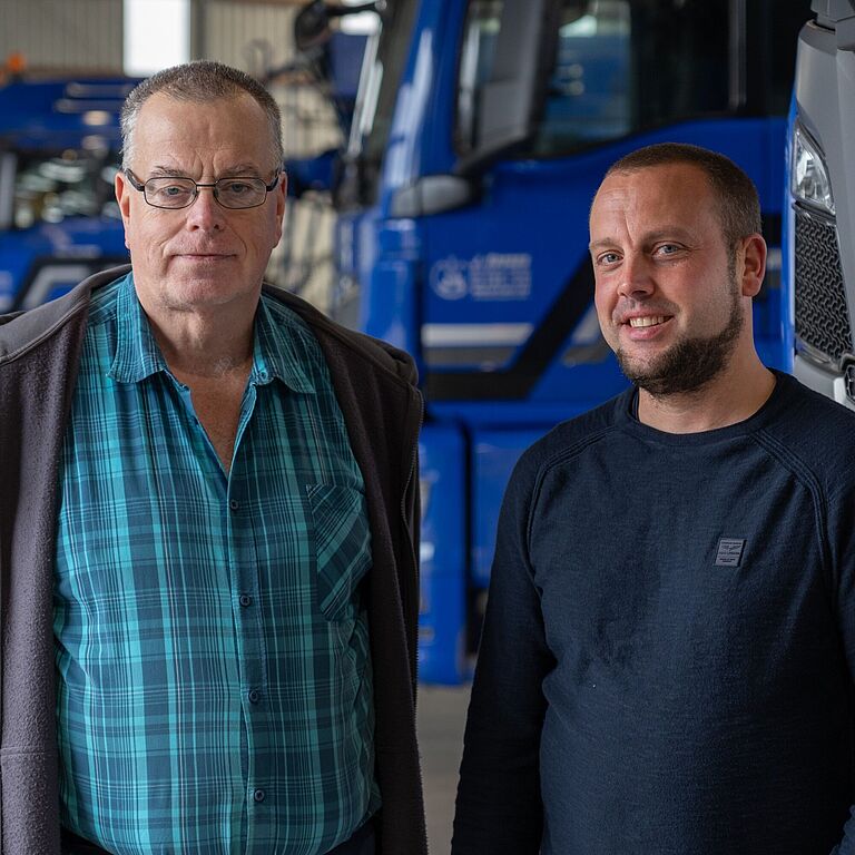 Zwei Männer stehen nebeneinander in einer Fahrzeughalle und schauen in die Kamera. Im Hintergrund stehen parkende Lkw.
