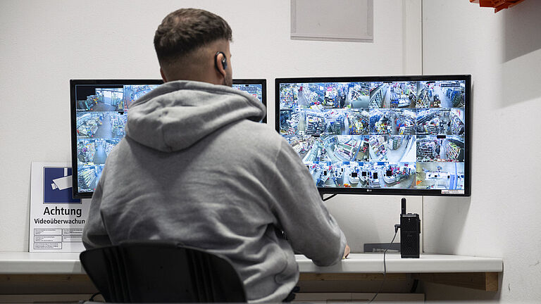 Ein Mann im grauen Kapuzenpulli sitzt vor zwei Bildschirmen und sieht die Bilder einer Videokamera.