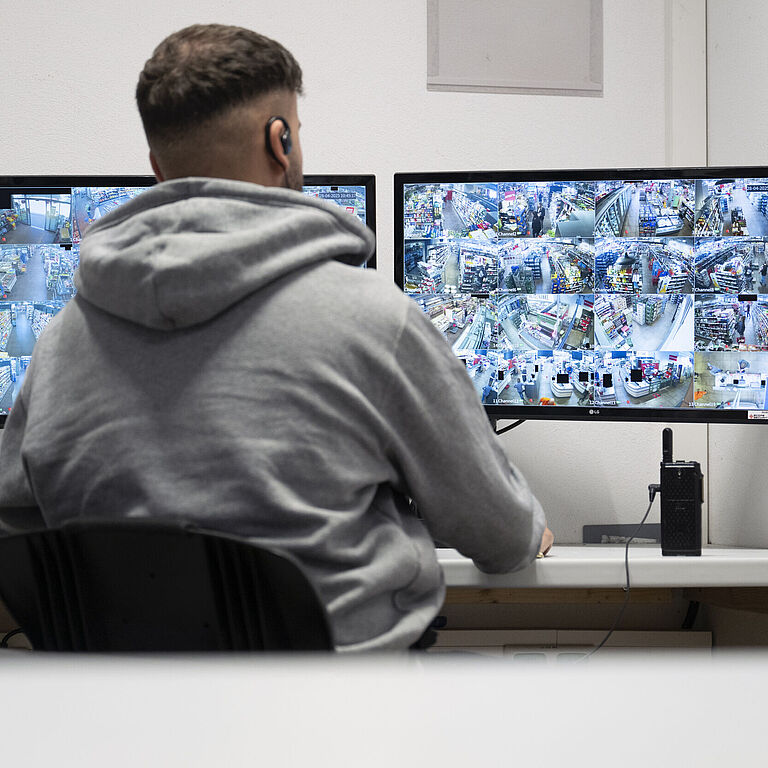 Ein Mann im grauen Kapuzenpulli sitzt vor zwei Bildschirmen und sieht die Bilder einer Videokamera. 
