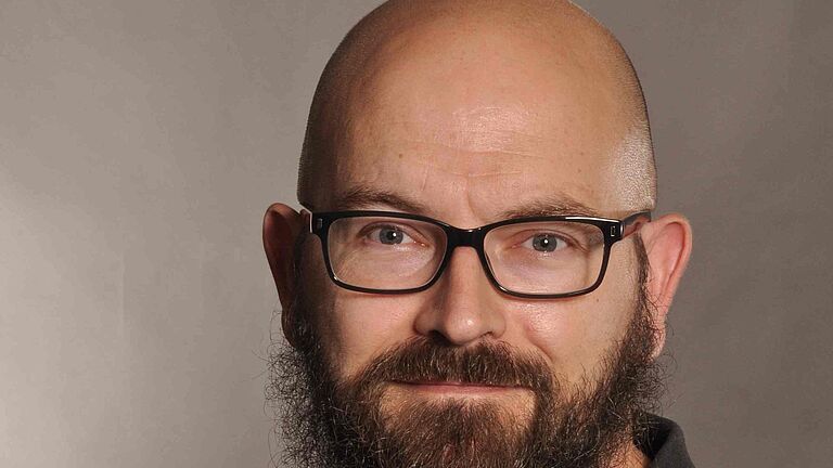 Ein Porträtfoto vor braun verlaufendem Hintergrund: Ein Mann mit Vollbart, Brille und Glatze, schaut freundlich in die Kamera. Er trägt ein schwarzes Hemd. 