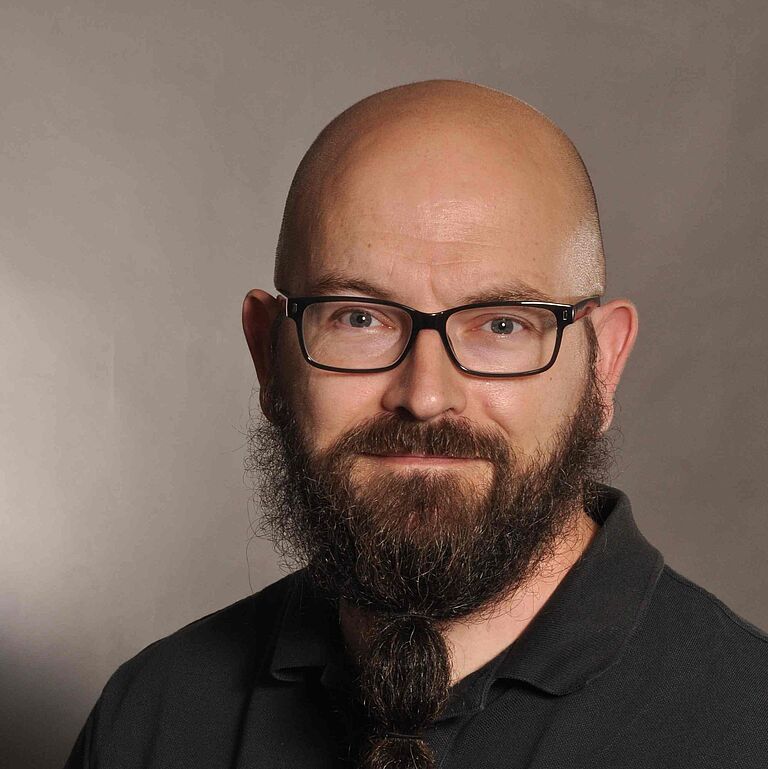 Ein Porträtfoto vor braun verlaufendem Hintergrund: Ein Mann mit Vollbart, Brille und Glatze, schaut freundlich in die Kamera. Er trägt ein schwarzes Hemd. 