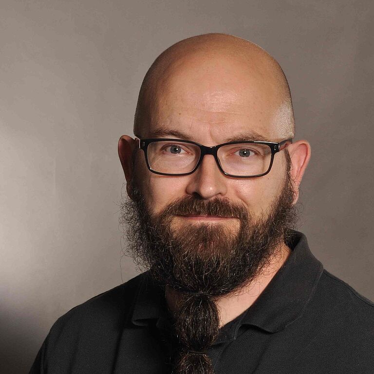 Ein Porträtfoto vor braun verlaufendem Hintergrund: Ein Mann mit Vollbart, Brille und Glatze, schaut freundlich in die Kamera. Er trägt ein schwarzes Hemd. 