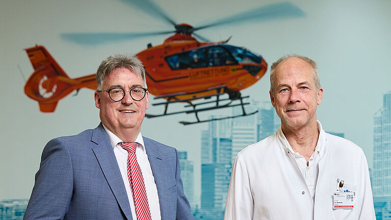 BGHW-Referent Martin Kögler, links stehend im blauen Anzug und Dr. Christoph Reimertz, Direktor der BG-Reha-Klinik, rechts im weißen Kittel, schauen freundlich in die Kamera. im Hintergrund ist das Bild eines Rettungshubschraubers.